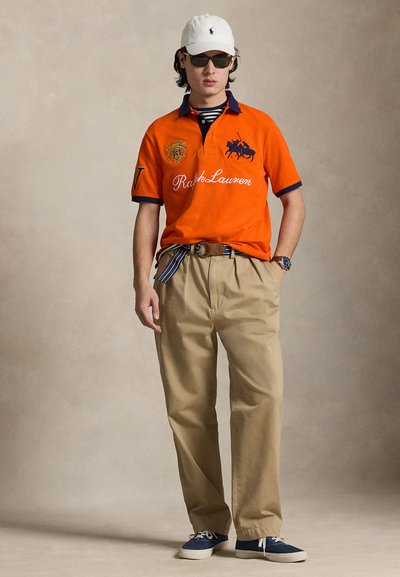 Πορτοκαλί πόλο μπλούζα με ναυτικές λεπτομέρειες, κεντημένο έμβλημα και το κείμενο «Ralph Lauren»· μπεζ παντελόνια· άσπρο καπέλο· ναυτικά αθλητικά παπούτσια· γυαλιά ηλίου.