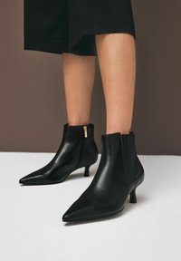 Černé kožené kotníkové boty s špičatou špičkou, nízkým podpatkem a zipem na boku. Mají hladkou texturu a elastický panel.
