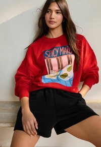Rød sweatshirt med et grafisk design, der viser drikkevarer på et bord. Kombineret med sorte shorts, der viser en afslappet pasform og blød tekstur.