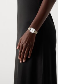 Montre carrée avec un cadran blanc, bracelet en métal argenté et doré, équipée de fines aiguilles des heures et des minutes, portée sur une main contre une robe noire.