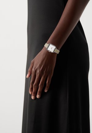 Montre carrée avec un cadran blanc, bracelet en métal argenté et doré, équipée de fines aiguilles des heures et des minutes, portée sur une main contre une robe noire.