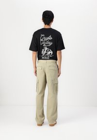 Camiseta negra con gráfico blanco "Crypto Cowboy" en la parte posterior, combinada con pantalones cargo beige que cuentan con múltiples bolsillos y un corte holgado.