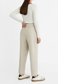 Žena s dlouhými hnědými vlasy, oblečená v bílém žebrovaném topu s dlouhým rukávem, béžových kalhotách s vysokým pasem a bílých teniskách s černými detaily.