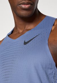 Modrá športová tielko s perforovanou textúrou a čiernym logom Nike. Má hlboký výstrih a bezrukávový dizajn pre lepšiu ventiláciu.