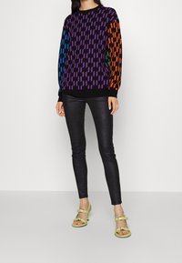 Maglione a maglia multicolore con motivi geometrici in viola, blu, arancione e verde su uno sfondo nero; indossato con leggings neri lucidi e sandali gialli.