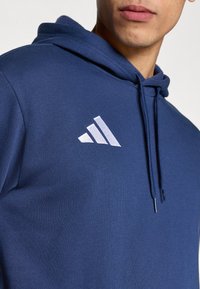 Μπλε σκούρο φούτερ από μαλακό ύφασμα, με τσέπη  μπροστά, κουκούλα με κορδόνι και λευκό λογότυπο με τρεις ρίγες στο στήθος.