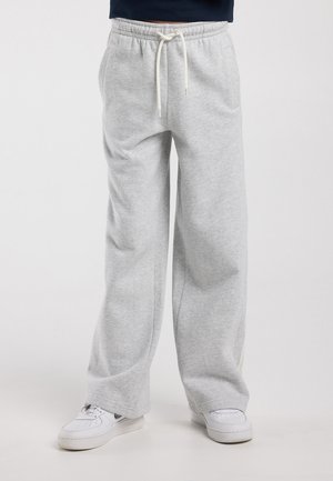 Pantalones deportivos de pierna ancha en gris claro con cintura elástica y cordón blanco, usados con zapatillas blancas sobre fondo neutro.