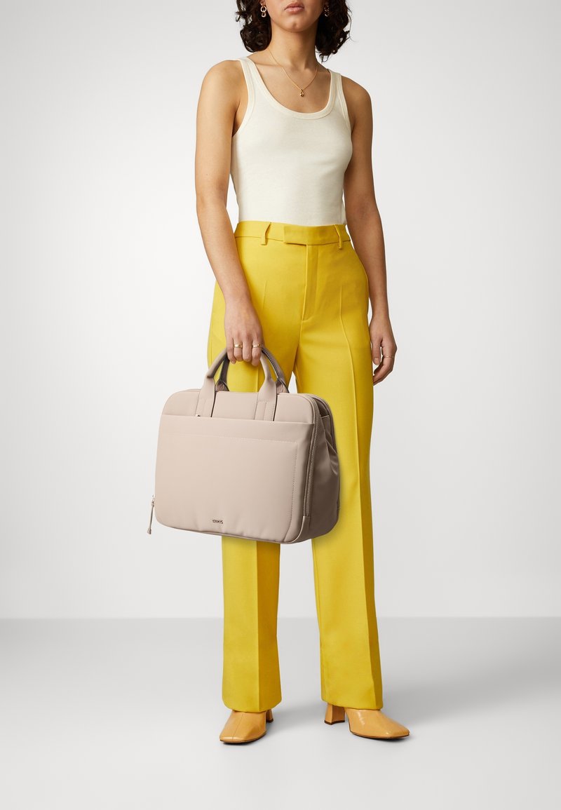 Kvinde iført hvid tanktop, gule bukser og beige sko, der holder en beige taske i læderstil med håndtag ved sin side.