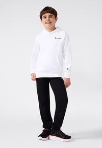 Sudadera blanca con capucha y bolsillo frontal, que presenta un pequeño logo negro. Pantalones de chándal negros y zapatillas negras con acentos de color completan el atuendo.
