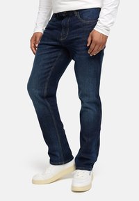 Dunkelblaue Jeans aus Denim mit geradem Schnitt, kontrastierenden Nähten und einem klassischen Fünf-Taschen-Design. Kombiniert mit weißen Sneakers.
