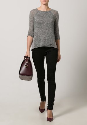 Žena v šedém svetru a černých kalhotách, držící tmavě bordó kabelku a nosící odpovídající bordó vysoké podpatky.