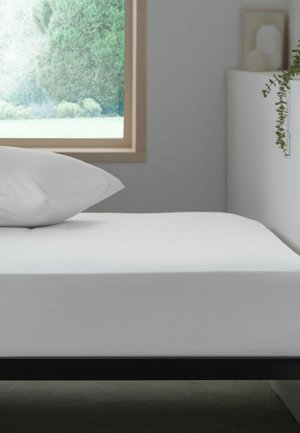 Minimalistisches weißes Bett mit weißem Kissen neben einem Fenster, das grüne Bäume zeigt, und einem weißen Regal mit einer Pflanze in einem hellen Raum.