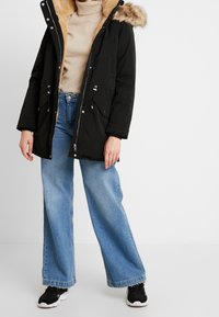Parka noire avec capuche en fausse fourrure, fermeture éclair à l'avant et détails à cordon. Portée sur un pull beige et un jean large bleu clair, associée à des baskets noires.