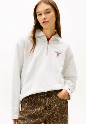 Grauer Halbreißverschluss-Sweatshirt mit roter Stickerei, kombiniert mit braunen Jeans im Leopardenmuster mit Seitentaschen und sichtbarem Logo-Tag.
