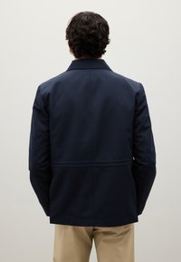 Mann mit dunklem, lockigem Haar, der eine marineblaue Jacke und beige Hosen trägt, steht vor einer schlichten, hellen Wand.