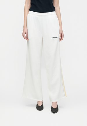 Donna che indossa pantaloni sportivi a gamba larga bianchi con il logo Calvin Klein sulla coscia, abbinati a scarpe nere a punta e a un top nero.