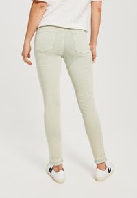 Jeans slim de color verde claro con bolsillos traseros, dobladillo deshilachado y textura sutil, combinados con zapatos blancos con acentos negros.