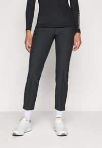 Mulher vestindo calças pretas justas, meias brancas até o tornozelo, tênis brancos e uma blusa preta de mangas longas com texto nas mangas, encostada contra um fundo simples.