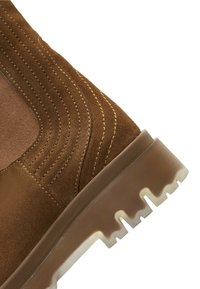 Bottine en suède marron avec des panneaux élastiques sur les côtés, couture texturée et une semelle en caoutchouc blanc robuste avec un motif dentelé.
