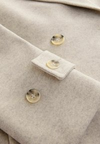 Tejido de lana beige con una textura cepillada, que presenta dos grandes botones translúcidos y una pestaña a juego. Diseño simple y limpio.