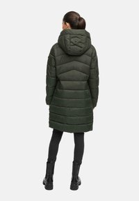 Vrouw met een donkere paardenstaart, gekleed in een lange olijfgroene gewatteerde pufferjas met capuchon, zwarte leggings en zwarte enkellaarsjes, staande met de rug naar de camera gekeerd.
