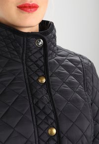 Quilted black jacket featuring a high collar, large gold buttons, and contrasting stitching, with a smooth, glossy texture.