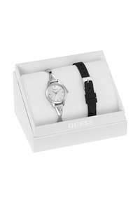 Montre-bracelet en argent de marque Guess avec un cadran blanc et un bracelet en métal, accompagnée d'un bracelet en silicone noir de remplacement, le tout dans une boîte de présentation blanche.