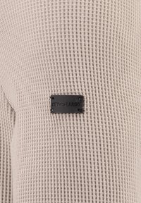 Beige strukturierter Stoff mit einem Rautenmuster, versehen mit einem schwarzen rechteckigen Etikett, auf dem "KEY LARGO" geprägt ist und an der Seite befestigt ist.