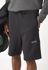 Črne atletske hlače s pasom na zavezo, ki imajo stranske žepke in svetlo zeleni logotip "BOSS". Gladka tkanina z rahlim sijajem.