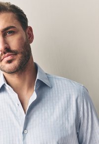 Uomo con capelli castani corti e barba che indossa una camicia a quadri azzurra chiara davanti a una parete beige uniforme.