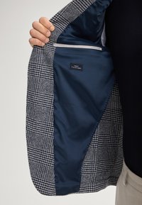 Blazer con tessuto esterno a quadretti in blu navy e bianco. L'interno presenta una fodera liscia blu navy e un'etichetta che indica "Miglior Tessuto Italiano."