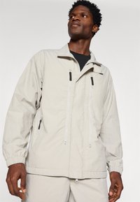 Giacca a vento grigia chiara con polsini elastici, due tasche frontali con zip e colletto alto. Il tessuto sembra leggero e texture.