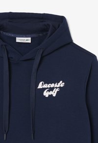 Felpa navy blue realizzata in tessuto morbido, con il logo bianco "Lacoste Golf". Progettata con cappuccio regolabile e vestibilità comoda.