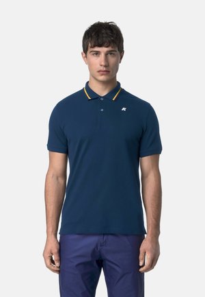 Giovane uomo in piedi, rivolto in avanti, indossa una polo blu navy con colletto a righe gialle e pantaloni blu scuro su sfondo semplice.
