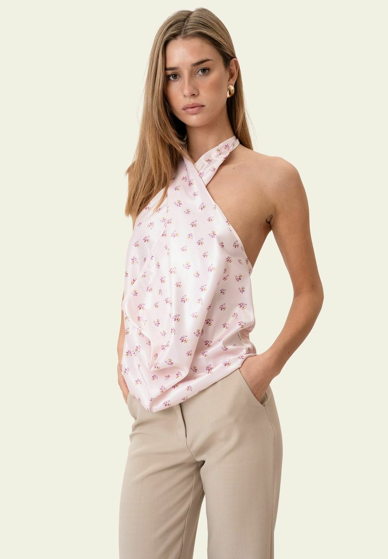 Femme aux cheveux bruns clairs et raides, portant un haut licou sans manches rose pâle à motif floral et un pantalon beige, debout les mains dans les poches.