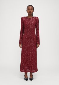 Raudona sequininių blizgučių suknelė ilgomisRankovėmis, suapvalintu kaklu, prigludusi siluetas ir tiesi sijonas, dėvima su juodais smailiais aukščiais.