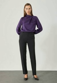 Blouse en satin violet avec détail de lien sur le côté, associée à un pantalon noir cintré et des talons noirs. Texture lisse et silhouette épurée.