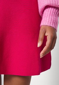 Vestido de punto fucsia con textura acanalada, que cuenta con una manga de punto en suave tono rosa. Primer plano de una mano descansando cerca del dobladillo, destacando los materiales.