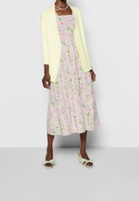 Ljusgul kofta över en blommönstrad rosa klänning med gröna accenter. Klänningen har en lagerdesign och en rufsig halsringning. Ljusgröna skor.