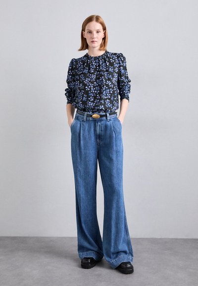 Schwarze Bluse mit Blumenmuster und Puffärmeln, kombiniert mit weiten Jeanshosen. Auffällige goldene Gürtelschnalle und schwarze klobige Schuhe.