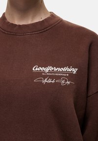 Felpa marrone con colletto a costine, caratterizzata da testo stampato bianco: "Goodfornothing" e "Tutti i diritti riservati" con accenti in firma.