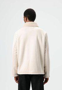 Beige jas met een zachte, gestructureerde kraag, rechte snit en zichtbare stikseldetails. De achterkant heeft een glad en strak ontwerp.