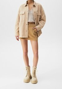 Veste chemise oversize beige, top court en maille blanche, jupe en denim beige effilochée, et bottes de combat crème avec semelles épaisses.