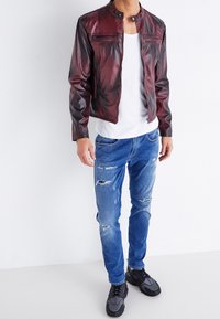 Chaqueta de cuero burdeos con cuello alzado, cierre frontal de cremallera y dos bolsillos con cremallera, combinada con vaqueros azul desgastados y zapatillas negras.