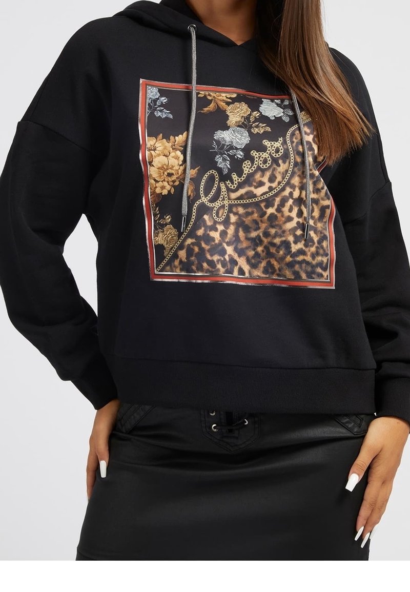 Femme porte un sweat à capuche noir avec un carré imprimé floral et léopard, texte "Guess" en motif chaîne, associé à une jupe en cuir noire et de longs ongles.