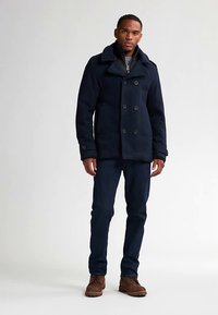 Cappotto doppiopetto blu navy con grandi bottoni, revers strutturati e tessuto testurizzato. Abbinato a jeans scuri e stivali marroni.