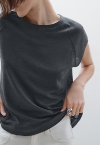 T-shirt leggero di colore grigio scuro con scollatura rotonda e maniche a cappuccio. Il tessuto testurizzato si adagia dolcemente sul corpo. Abbinato a pantaloni bianchi.