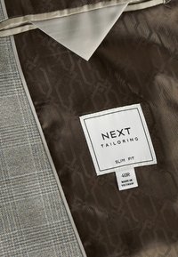 Grau- und braun kariertes Gewebe mit einem glatten Innenfutter. Das Etikett trägt die Aufschrift "NEXT TAILORING", welches auf eine Slim-Fit-Passform, Größe 40R, hergestellt in Vietnam hinweist.