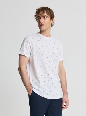 Giovane uomo con maglietta bianca a maniche corte con piccole icone nere stampate, in piedi con una mano in tasca, che guarda leggermente verso sinistra.