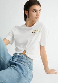 T-shirt en coton blanc avec un détail de nœud circulaire à la taille, impression du logo doré, associé à un jean taille haute bleu clair.